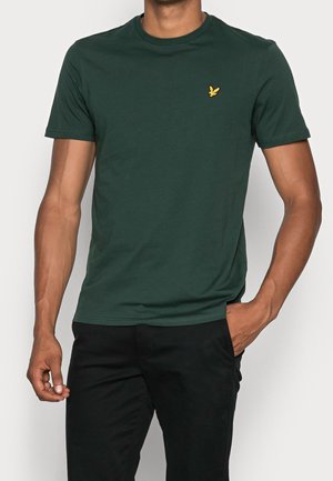 Man draagt een donkergroen T-shirt met korte mouwen en een klein geel vogellogo op de borst, zwarte broek, één hand in de broekzak, neutrale achtergrond.