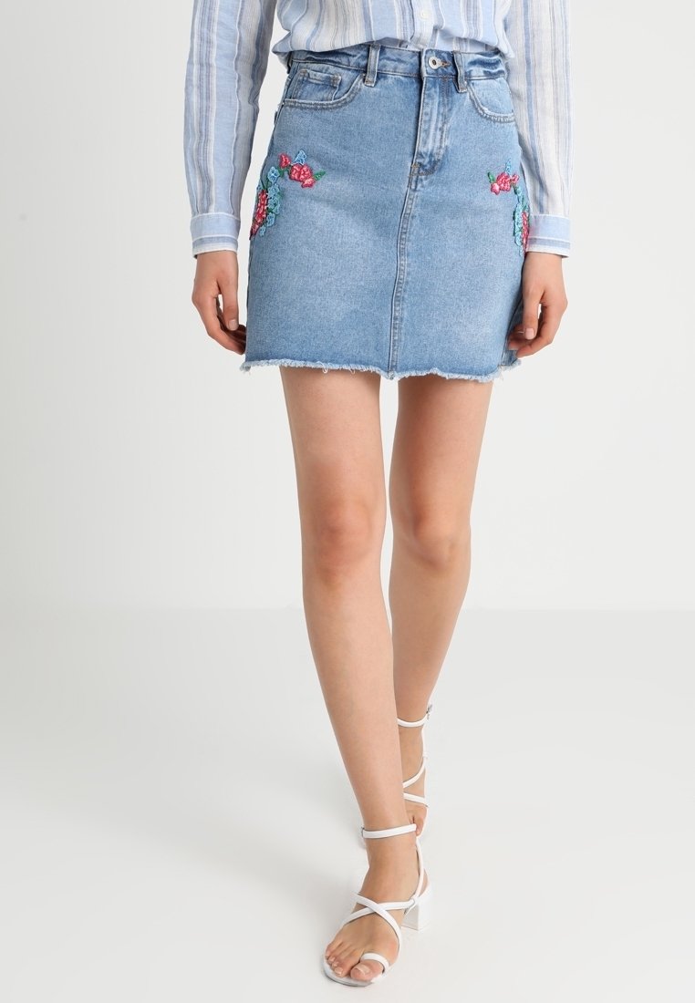 Minigonna in denim con orlo sfilacciato. Caratterizzata da ricami floreali colorati su entrambi i lati. Indossata con sandali con cinturini di colore chiaro.