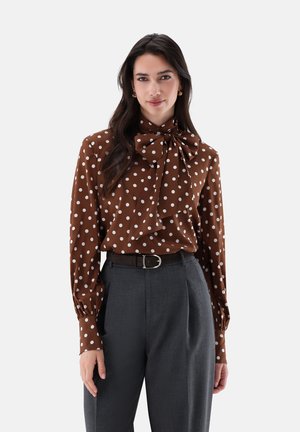 Frau mit langen dunklen Haaren, die eine braune gepunktete Bluse mit großer Schleife und hochtaillierte dunkelgraue Hose mit Gürtel trägt.