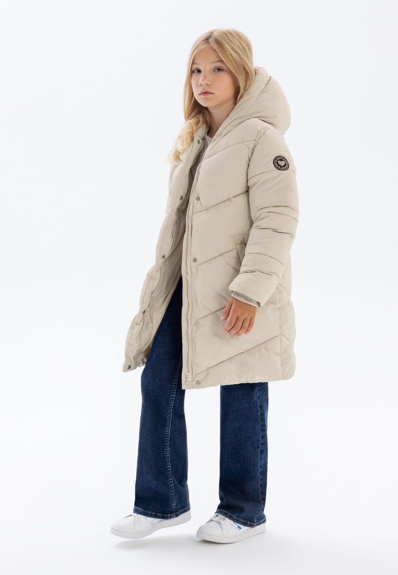 Doudoune beige avec capuche, présentant un quilting diagonal et un patch logo circulaire sur la partie supérieure de la manche. Associée à un jean en denim et des chaussures blanches.