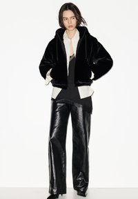 Černá krátká kapucová mikina z umělé kožešiny se šňůrkami, vrstvená přes bílé pruhované tričko a černý krajkový top, doplněná lesklými černými koženými kalhotami.