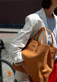 Sac en cuir tanné avec des poignées roulées, des accents de grommet et une base structurée. Accompagné d'un blazer de couleur claire et d'une chemise grise.