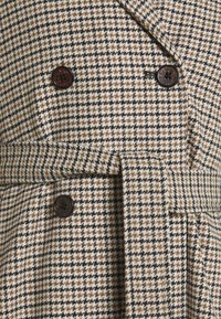 Dvojradový kabát v vzore houndstooth, ktorý obsahuje hnedé, čierne a krémové farby. Obsahuje tmavé gombíky a opasok v páse.