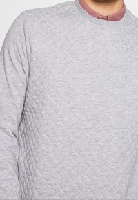 Sweat-shirt gris matelassé avec un col rond et un motif texturé. En dessous, une chemise à col dans une couleur neutre complémentaire.