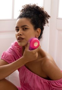 Circular facial cleansing device in bright pink with textured surface and gold logo, held against skin, set near a window with soft light.