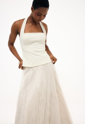 Kvinde iført en hvid ærmeløs haltertop og en lang hvid plisseret nederdel, kigger nedad med hænder, der holder siderne af hendes top.