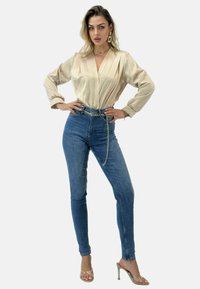 Beige satinbluse med pliser, højtaljede blå jeans, kædebeltdetalje og gennemsigtige højhælede sko, stående på en hvid baggrund.