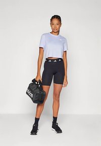 Noor naine, kes kannab lilla lühikest T-särki ja musti Adidas lühikesi pükse, hoiab musta Adidas spordikotist seistes tasase taustaga.