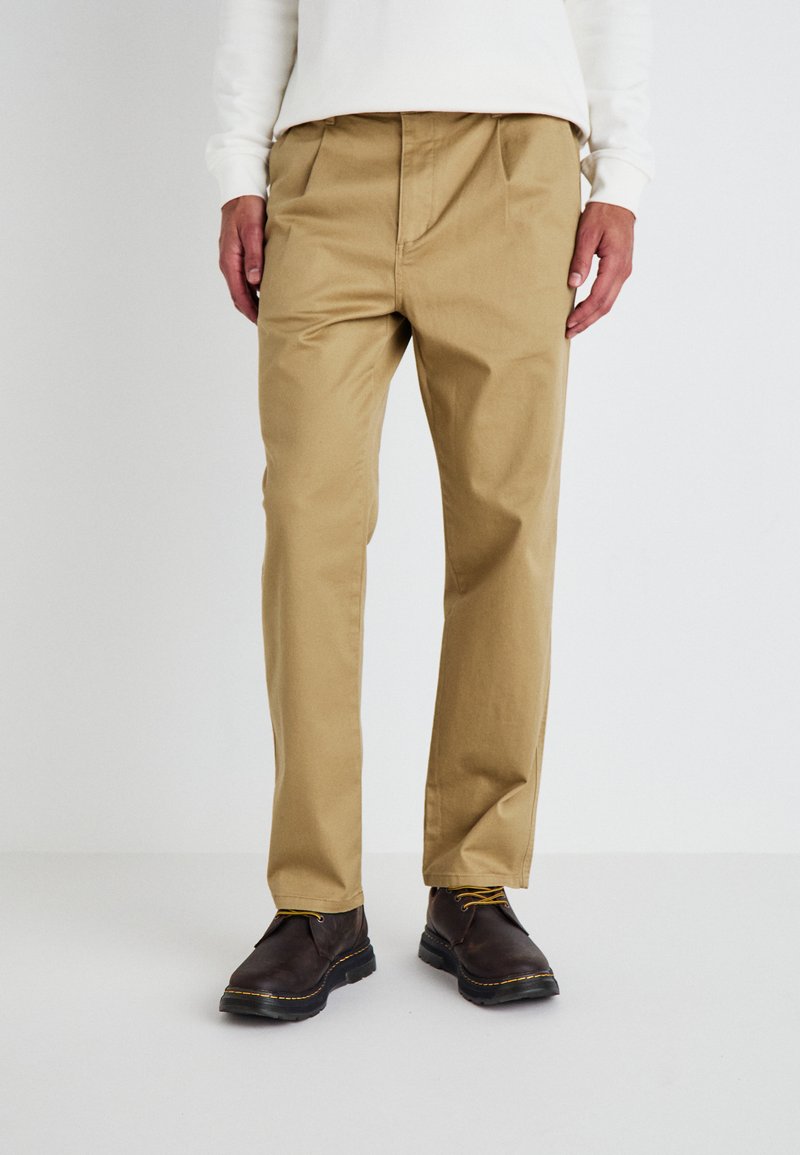 Khaki pants made of cotton blend fabric with a straight design, featuring front pleats. Paired with brown leather shoes.