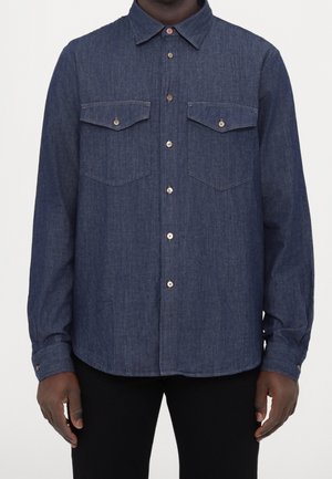 Mand iført en langærmet mørkeblå denimskjorte med knappede brystlommer og beige knapper, kombineret med sorte bukser.