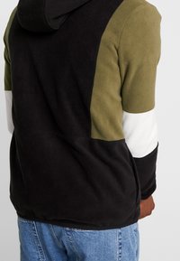 Felpa con cappuccio in fleece, caratterizzata da pannelli neri, verde oliva, bianchi e crema. Include un cappuccio e presenta una finitura morbida e lavorata.