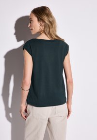 Tmavě zelený krátký rukáv top vyrobený z měkké tkaniny, s kulatým výstřihem a jemnými švy, doplněný světlými kalhotami.