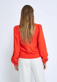 Red, puff-sleeve sweater with a fitted waist. Soft fabric, smooth texture, and ribbed cuffs. Worn with light-colored trousers.