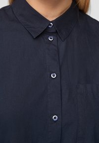 Marinblå knäppskjorta med spetsig krage, med matchande knappar och en liten ficka på vänster sida. Slät tygtextur.