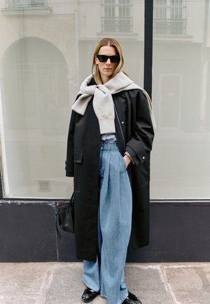 Femme portant un manteau noir oversize, un sweat-shirt gris drapé sur les épaules, un jean bleu large, des lunettes de soleil noires et tenant un sac à main noir, debout à l'extérieur.