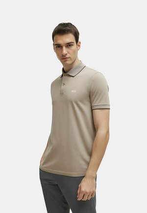 Uomo che indossa una polo beige a maniche corte con rifiniture e bottoni neri, in piedi con una mano in tasca davanti a uno sfondo semplice.