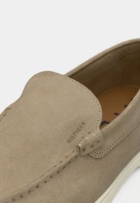 Beige suede slip-on shoe with a stitched moccasin design, featuring a smooth texture and white rubber sole. "HILFIGER" logo on side.