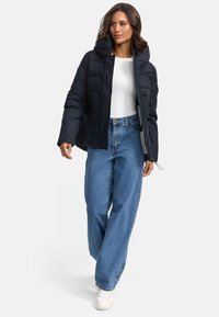 Puffig svart jacka med huva, med dragkedjestängning och sidofickor, bärs över en vit topp och lösa blå jeans.