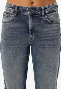 Džínsy s vysokým pásom vyrobené z vyblednutého modrého denimu. Majú zapínanie na gombík, predné vrecká a viditeľné švy.