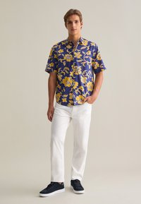 Camisa de manga corta en azul oscuro con un gran patrón floral amarillo, diseño abotonado, llevada con pantalones blancos y zapatillas azul marino.