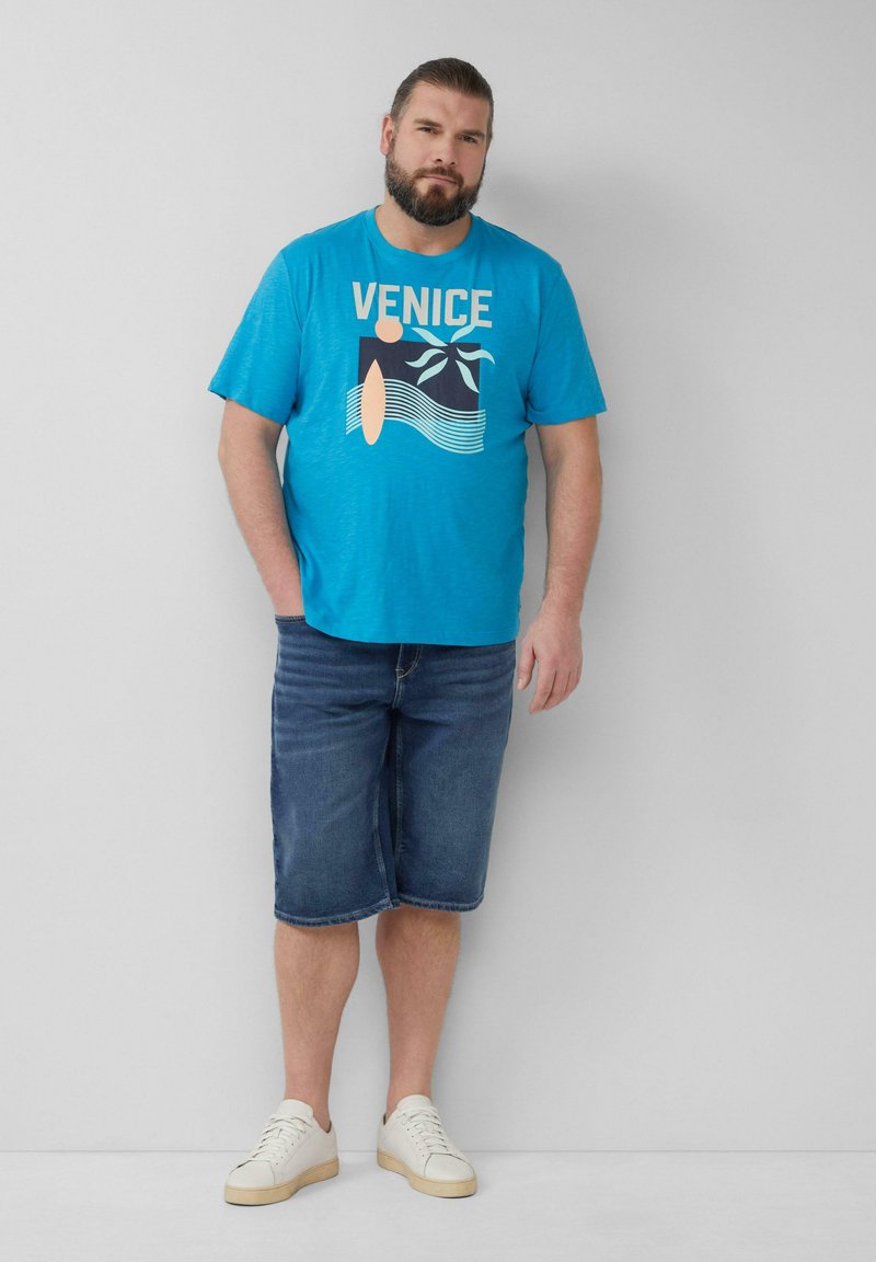Blauwe t-shirt met "VENICE" afbeelding, gedragen met knielange denimshorts en witte sneakers. Effen achtergrond.