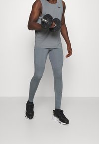 Mann in grauem Sportoberteil und Leggings hebt eine schwarze Kurzhantel und steht auf einem weißen Boden vor einer weißen Wand.