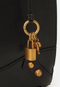 Sac en cuir noir avec des accessoires en métal doré, y compris une bague, un cadenas et des clés. Présente des coutures décoratives et des clous en métal.