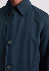 Marineblauer Button-Down-Mantel mit spitzem Kragen und drei sichtbaren Knöpfen, getragen über dem Oberkörper einer Person vor einem schlichten Hintergrund.