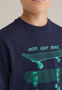 Marineblå sweatshirt med rund hals som har en grön grafisk design av skateboardar och texten "SKATE, SLEEP, REPEAT." Mjuk bomullsmaterial.