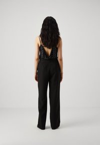 Kobieta z długimi ciemnymi włosami, ubrana w czarną bluzkę bez rękawów ozdobioną cekinami z odkrytymi plecami oraz w wysokie czarne szerokie spodnie, stojąca przodem do widza.