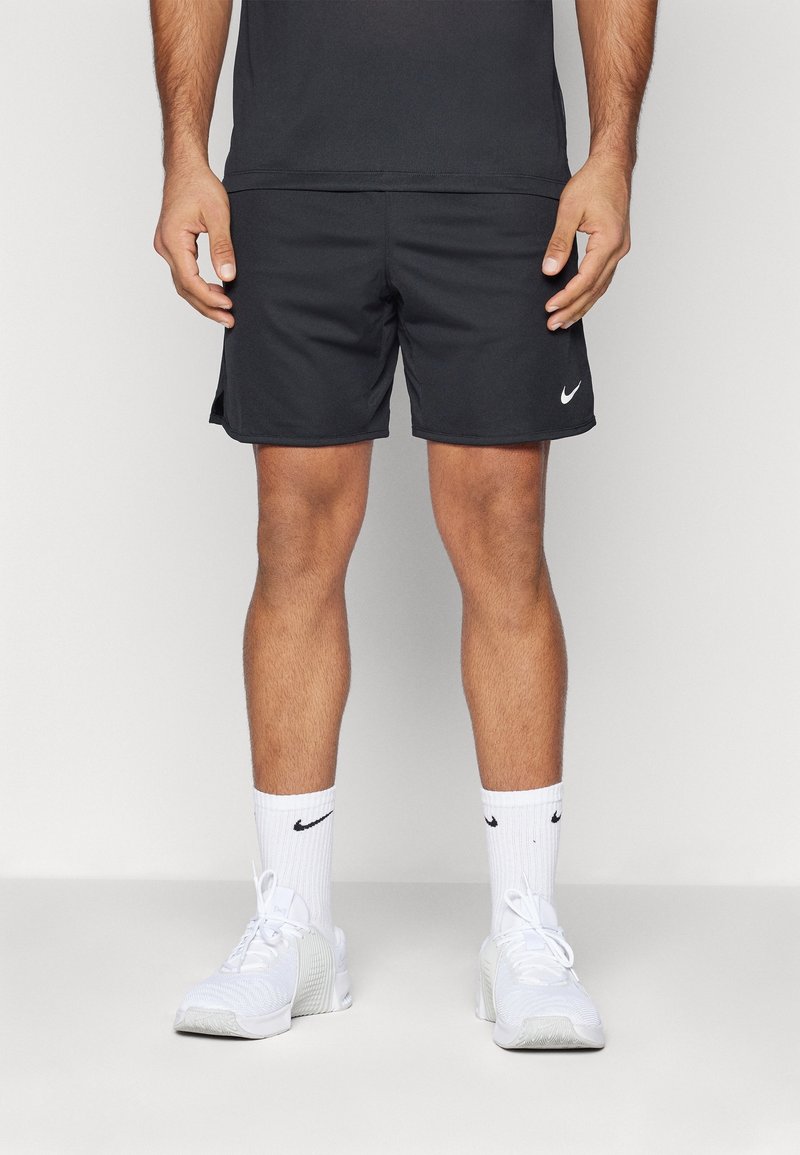 Homme portant un short de sport noir Nike, des chaussettes blanches Nike et des chaussures de sport blanches, debout sur un fond clair uni.