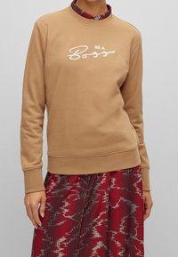 Sudadera beige con cuello redondo y mangas largas. Presenta un texto bordado en blanco que dice "BE A Boss." Llevada sobre una falda roja con estampado.