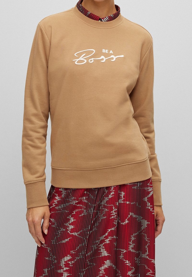 Sudadera beige con cuello redondo y mangas largas. Presenta un texto bordado en blanco que dice "BE A Boss." Llevada sobre una falda roja con estampado.