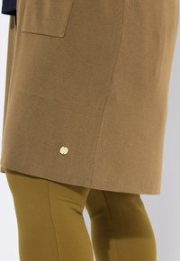 Braunes Strickkleid mit Saum und Seitentaschen über olivgrünen Leggings, getragen von einer Person, die vor einem weißen Hintergrund steht.