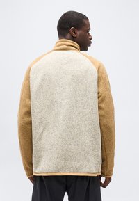 Άνδρας που φαίνεται από π ίσω, φοράει μπεζ και ανοιχτό γκρι φλις μπουφάν με ψηλό γιακά, μπροστά σε απλό φόντο.