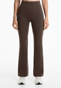Leggings marron évasés en tissu extensible avec taille haute. Texture lisse, coutures minimales et forme ajustée. Portés avec des chaussures de sport blanches.