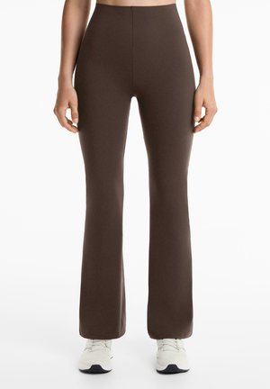 Leggings marron évasés en tissu extensible avec taille haute. Texture lisse, coutures minimales et forme ajustée. Portés avec des chaussures de sport blanches.