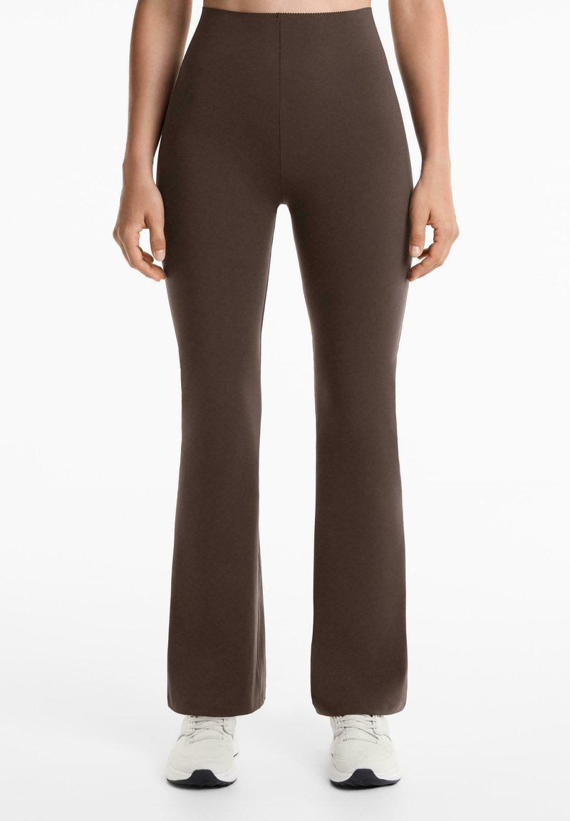 Leggings marron évasés en tissu extensible avec taille haute. Texture lisse, coutures minimales et forme ajustée. Portés avec des chaussures de sport blanches.