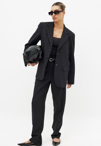 Črn krojen blazer z velikimi kragnami, v kombinaciji s črno majico brez rokavov, širokimi hlačami, elegančnimi sončnimi očali in geometrijsko črno torbico.