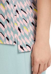 Primer plano de una mano reposando junto a una cadera, vistiendo una camisa estampada en rosa, azul verdoso, negro y amarillo, con pantalones azul claro.