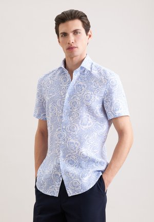 Młody mężczyzna z ciemnymi włosami, ubrany w jasnoniebieską i białą koszulę w kwiaty z krótkim rękawem oraz ciemne spodnie, stoi z rękami w kieszeniach.