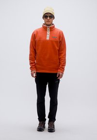 Πορτοκαλί φλις μπλούζα με λευκό γιακά με κουμπιά, φορεμένη με μαύρο παντελόνι και γκρι ορειβατικά μποτάκια. Αξεσουάρ περιλαμβάνουν γυαλιά ηλίου και μπεζ σκούφο.