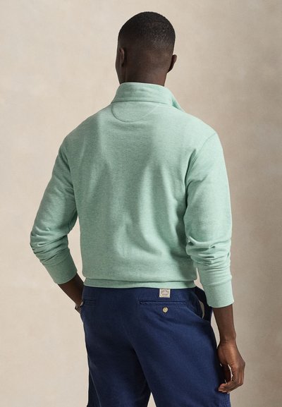 Sweatshirt verde menta com gola alta, punhos e bainha canelados. Usado com calças azul-marinho com bolsos traseiros e um pequeno detalhe de etiqueta.