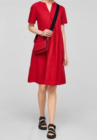 Rød kjole med korte ærmer, V-hals, samlet talje og en glat tekstur. Sammen med en matchende rød crossbody taske og sorte sandaler.