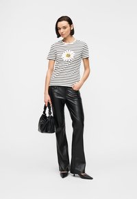 Γυναίκα που στέκεται φορώντας μια λευκή μαύρη ριγέ μπλούζα με ένα μεγάλο μαργαρίτα, μαύρο δερμάτινο παντελόνι, μυτερά τακούνια και κρατώντας μια μαύρη τσάντα.