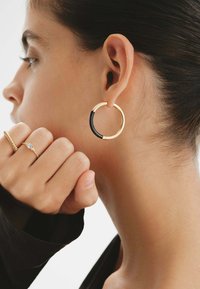 Pendientes de aro dorados con un acento negro, que presentan una textura suave y una forma circular, llevados en la oreja de una persona.