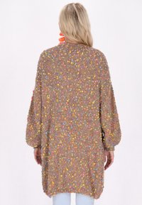 Stickad cardigan i beige med färgglada fläckar. Löst, oversized passform, sänkt axlar och vida ärmar. Mjuk textur och avslappnad design.