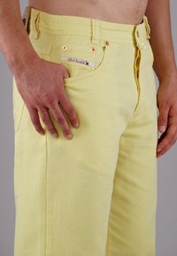 Yellow denim jeans with a smooth texture, featuring a front pocket and metal rivets. The waistband has a brand label and button closure.