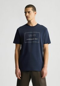 Tumesinine puuvillane T-särk, millel on ristkülikukujuline logo, kus on kontrastsete niitidega tekst "LIU-JO asutatud 1995". Lühikesed varrukad.