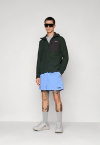 Tamsiai žalia užtraukiama vilnos striukė su pilku skydeliu, derinama su šviesiai mėlynais šortais. Pilkos kojinės ir balti sportbačiai užbaigia įvaizdį.